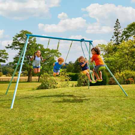 Four children play on the TP Toys TP Triple Metal Swing Set with Glider Swing Seat, which features adjustable seats. The blue swing set stands on grassy park grounds, surrounded by trees and greenery on a sunny day.