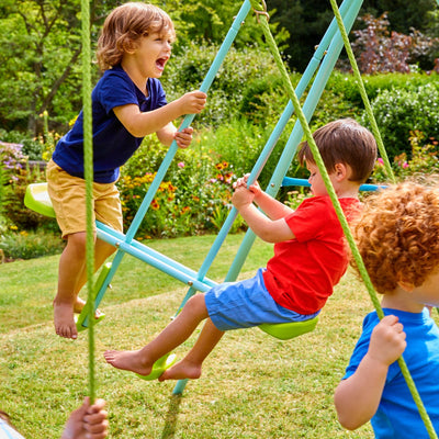 Three children play on the TP Toys TP Triple Metal Swing Set with Glider Swing Seat in a grassy garden, surrounded by colorful flowers and lush greenery. The adjustable swing seats make it perfect for active fun outdoors.
