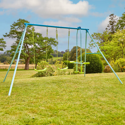 The TP Toys TP Triple Metal Swing Set with Glider Swing Seat sits on a grassy lawn, surrounded by trees and bushes on a clear day.