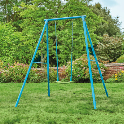 The TP Toys TP Deluxe Single Metal Swing Set, featuring a blue frame and adjustable seat, stands on a grassy lawn with green trees, shrubs, and a brick garden wall in the background.