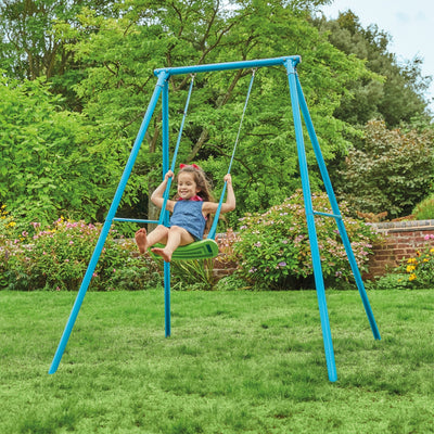 A young girl in a blue dress swings on the TP Toys TP Deluxe Single Metal Swing Set with an adjustable seat, surrounded by green grass and blooming flowers.