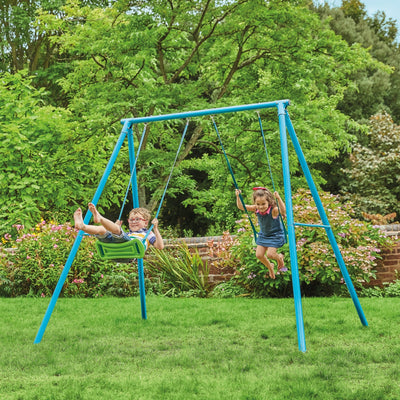 Two children play on the TP Toys TP Deluxe Double Metal Swing Set with Rapide swing seats and a blue powder-coated steel frame, set up in a grassy backyard surrounded by trees and flowering plants.