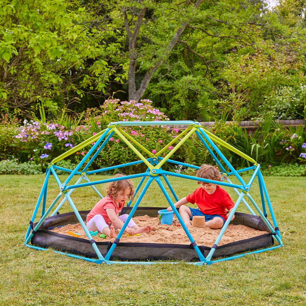Two young children play with sand toys inside the TP Toys 1.9m Metal Climbing Dome & Sandpit on a grassy lawn, surrounded by trees and flowers.