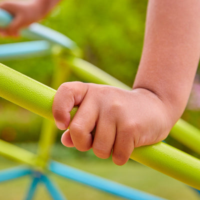A child grips a green metal bar on the TP Toys 1.9m Metal Climbing Dome & Sandpit, with blurred greenery in the background.
