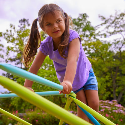 A young girl in a purple shirt and denim shorts climbs on the TP Toys 1.9m Metal Climbing Dome & Sandpit, surrounded by trees and flowers.