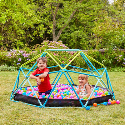 Two young children play on the TP Toys 1.9m Metal Climbing Dome & Sandpit, set on a garden lawn among trees and flowers—a perfect outdoor toy for active fun in any kids’ garden.