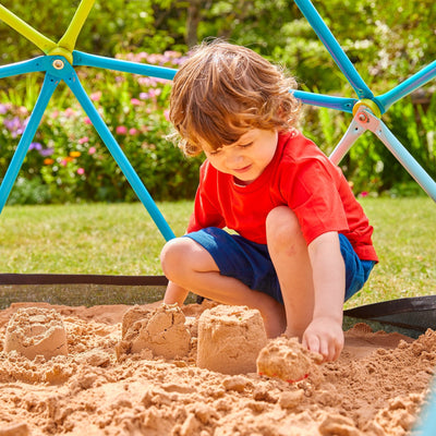 A child builds sandcastles in the TP Toys 1.9m Metal Climbing Dome & Sandpit, surrounded by greenery and outdoor play equipment, enjoying a fun-filled day with garden toys.