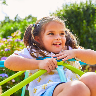 A young girl with pigtails smiles as she enjoys the TP Toys TP 1.9m Metal Climbing Dome & Sandpit, making the most of her outdoor playtime on a sunny day.