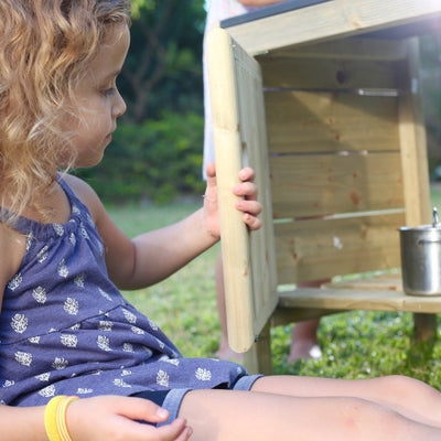 A young child outdoors opens a door on the TP Toys TP Junior Chef Wooden Mud Kitchen with Working Tap & Sink (FSC® certified), featuring a metal cup on top and greenery in the background.