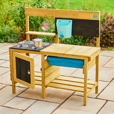 The TP Toys TP Deluxe Wooden Mud Kitchen - FSC® certified features a stove, sink, hanging utensils, chalkboard, storage shelves, and a blue bin. Set outdoors on a stone patio with garden plants in the background.