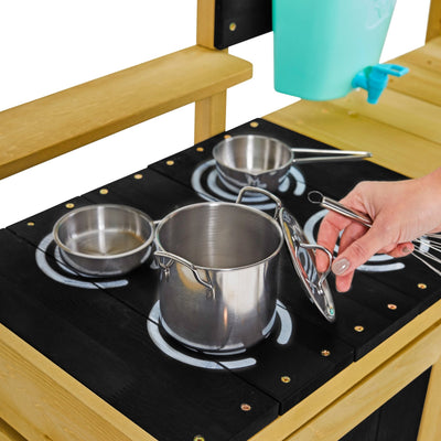 A hand holds a lid next to a stainless steel pot on a toy stove, with two metal pans nearby—ideal for imaginative play in the TP Deluxe Wooden Mud Kitchen by TP Toys, made from FSC® certified wood.