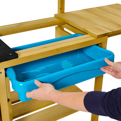 Someone places a blue plastic tray of water into the TP Deluxe Wooden Mud Kitchen by TP Toys, creating an ideal setup for creative outdoor play.