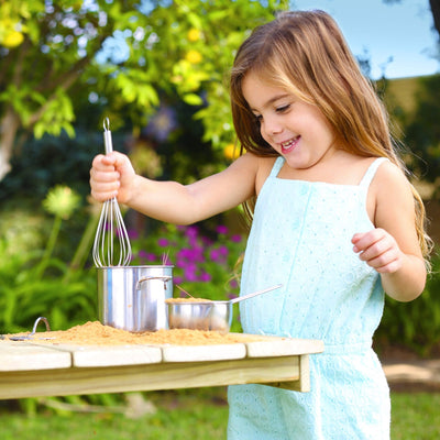 A young girl in a light blue dress uses a whisk and metal pot at the TP Toys TP Deluxe Wooden Mud Kitchen - FSC® certified, enjoying outdoor play on a sunny day.