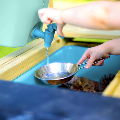 A child collects water from a blue tap into a metal pan at the TP Toys TP Deluxe Wooden Mud Kitchen - FSC® certified, with pinecones seen in a blue basin underneath.