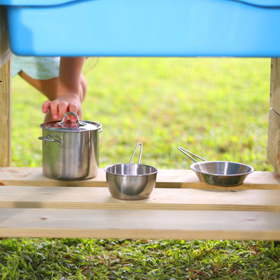 A child’s hand reaches for a lidded metal pot beside two measuring cups on the TP Toys TP Deluxe Wooden Mud Kitchen - FSC® certified, set outdoors with grass in the background.