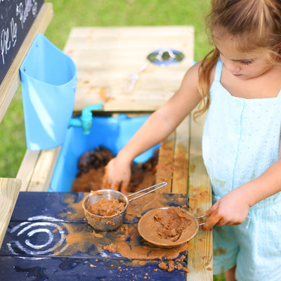 A young girl in a light blue outfit plays with wet sand at the TP Toys TP Deluxe Wooden Mud Kitchen – FSC® certified.
