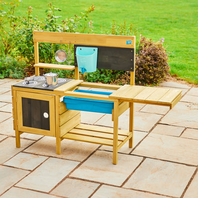 The TP Toys TP Deluxe Wooden Mud Kitchen - FSC® certified features stove burners, a sink, hanging utensils, a blue storage bin, and a fold-out side table—ideal for creative outdoor play.