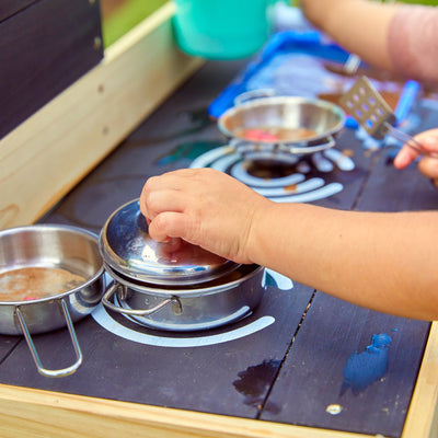 A child plays with metal pots, pans, and utensils on the TP Toys Little Chef Wooden Mud Kitchen - FSC® certified, enjoying outdoor fun and imaginative play.