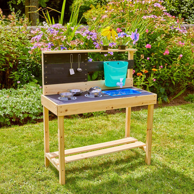 The TP Toys TP Little Chef Wooden Mud Kitchen - FSC® certified stands on grass with a blue sink, utensils, toy cookware, green bucket, and flowers—ideal for creative outdoor play in any garden.