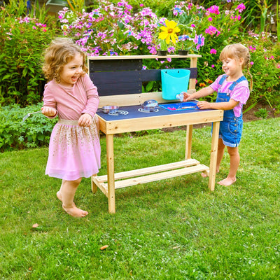 Two children enjoy outdoor play in the garden with the TP Toys TP Little Chef Wooden Mud Kitchen - FSC® certified, smiling as they use toy utensils, surrounded by flowers and greenery.