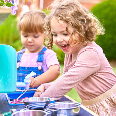 Two young children smile and play with water and kitchen utensils at the TP Toys TP Little Chef Wooden Mud Kitchen – FSC® certified, enjoying hands-on outdoor play.