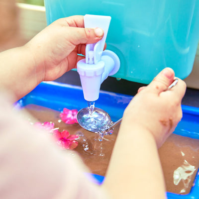A child explores with the TP Little Chef Wooden Mud Kitchen by TP Toys, scooping water from a blue dispenser with a metal spoon over a tray where pink objects float, inspiring curiosity and outdoor play.