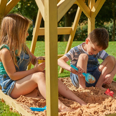 Two children play with sand toys in a wooden sandbox outdoors, beside the TP Toys Treehouse wooden play tower with panel kit, balcony, wavy slide & swing arm—FSC® certified—on a sunny day.