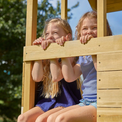 Two young girls sit side by side on a TP Toys Treehouse wooden play tower with panel kit, balcony, wavy slide & swing arm (FSC® certified), smiling and gazing out while resting their chins on their hands in the sunshine.