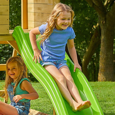 Two young girls enjoy the TP Toys Treehouse Wooden Play Tower, featuring a wavy slide, balcony, and panel kit—FSC® certified—for outdoor fun on a sunny day.