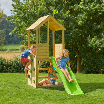 Three children play on the TP Toys Treehouse Wooden Play Tower, with a wavy slide and climbing wall (FSC® certified), in a grassy outdoor area surrounded by trees.