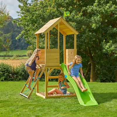 Three children play on the TP Toys Treehouse Wooden Play Tower, featuring a wavy slide, cargo net, and covered platform. The FSC® certified set is placed on grass with trees in the background.