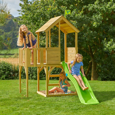 Three young girls play on the TP Toys Treehouse Wooden Play Tower, with Wavy Slide & Wooden Balcony (FSC® certified), set on a grassy lawn near trees.