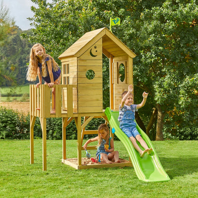Three children play on the TP Toys Treehouse Wooden Play Tower, featuring a wavy slide, wooden balcony, and panel kit (FSC® certified), set in a grassy outdoor area surrounded by trees.