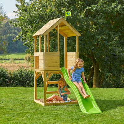 Two young girls play on the TP Toys TP Treehouse Wooden Play Tower with Wavy Slide—FSC® certified. One slides down the wavy slide while the other sits in a sandpit beneath the outdoor playhouse, surrounded by grass and trees.