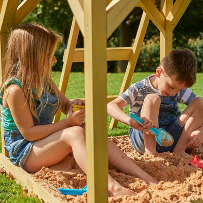 A boy and girl play in sand beside the TP Toys TP Treehouse Wooden Play Tower with Wavy Slide (FSC® certified), adding excitement to their outdoor adventures.