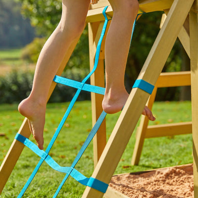 A child descends the TP Toys Treehouse Wooden Play Tower, with Wavy Slide & Cargo Net (FSC® certified), using a blue rope ladder above the sandbox on grass, while a wavy slide curves from the side, enhancing outdoor playtime.
