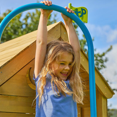 A young girl in a blue shirt smiles as she hangs from a blue bar in front of the TP Toys Treehouse Wooden Play Tower with Wavy Slide & Firemans Pole (FSC® certified) outdoors.