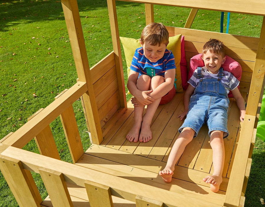 Two boys relax in the TP Toys Treehouse Wooden Play Tower—one sits with arms around his knees, the other leans on cushions. The FSC® certified tower features a wavy slide, wooden balcony, and basketball hoop.