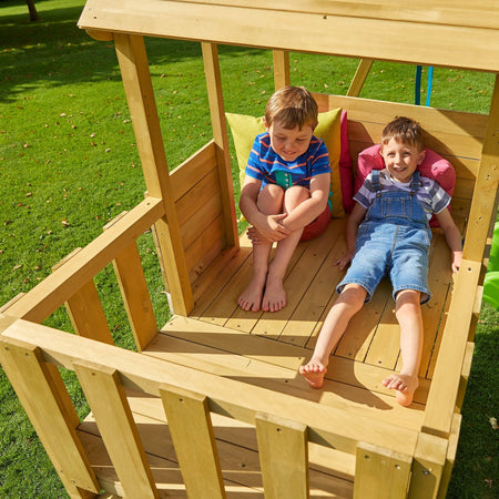 Two boys smile inside the TP Toys Treehouse Wooden Play Tower with Wavy Slide & Wooden Balcony (FSC® certified), one sitting cross-legged and the other reclining, both enjoying their cozy wooden playhouse on a grassy lawn.