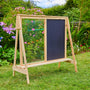 The TP Toys TP Two-Way Play Easel, with a clear acrylic panel on one side and a black chalkboard panel on the other, stands on grass surrounded by colorful flowers and greenery for creative play.