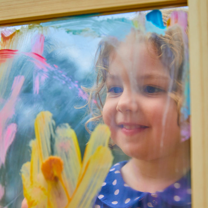 A young child in a blue polka dot shirt smiles while painting colorful patterns on the TP Toys TP Two-Way Play Easel, turning art time into creative play.