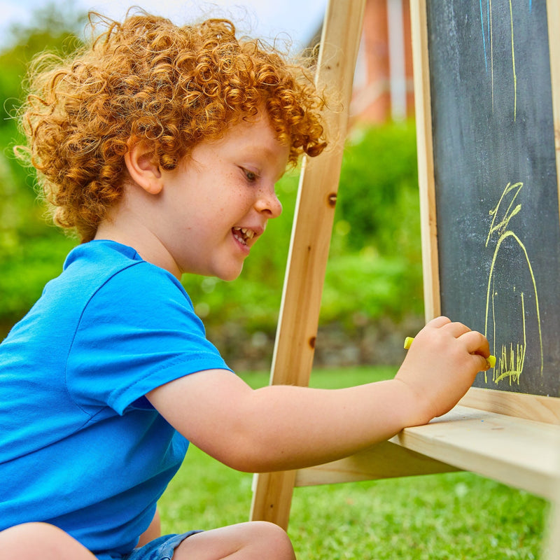 A young child with curly red hair enjoys creative play outdoors on a sunny day, drawing with yellow chalk on the blackboard side of the TP Toys TP Two-Way Play Easel.