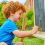A young child with curly red hair enjoys creative play outdoors on a sunny day, drawing with yellow chalk on the blackboard side of the TP Toys TP Two-Way Play Easel.