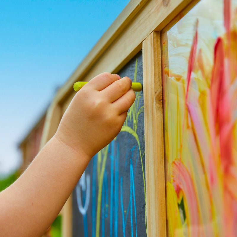 A child draws with yellow chalk on the TP Toys TP Two-Way Play Easel outdoors, creating colorful lines under a bright blue sky and sparking creative play.