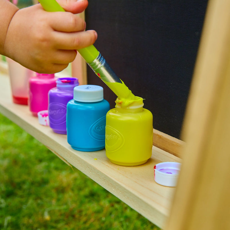 A hand dips a paintbrush into a yellow paint bottle, while blue, pink, and purple bottles sit nearby—great for creative fun with the TP Toys TP Two-Way Play Easel or any imaginative activity.