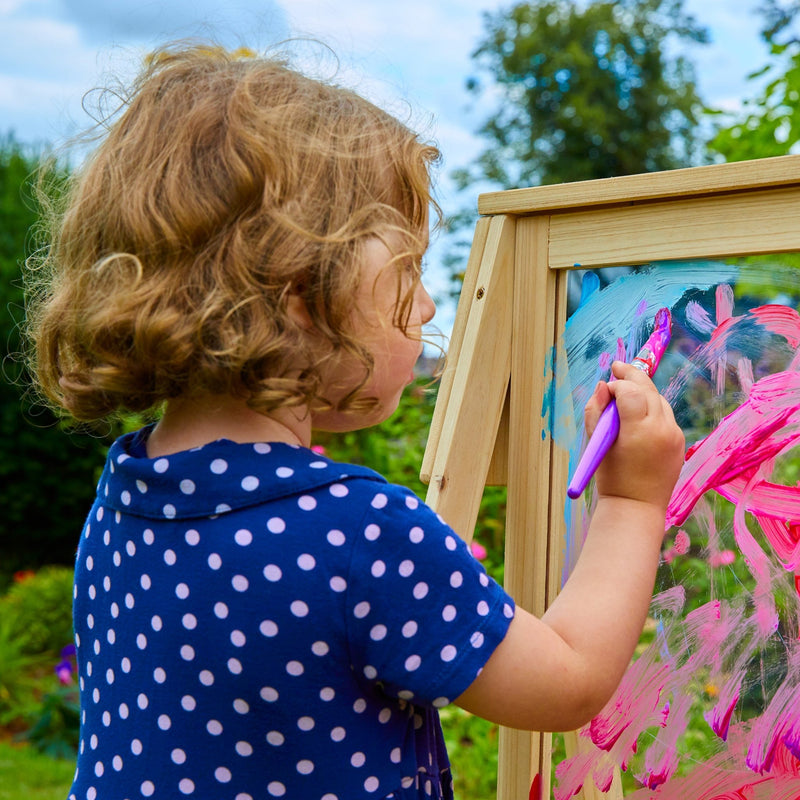 A young child with curly hair in a blue polka-dot shirt paints colorful shapes on the TP Toys TP Two-Way Play Easel outdoors, enjoying a creative toy that sparks imagination.