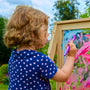 A young child with curly hair in a blue polka-dot shirt paints colorful shapes on the TP Toys TP Two-Way Play Easel outdoors, enjoying a creative toy that sparks imagination.