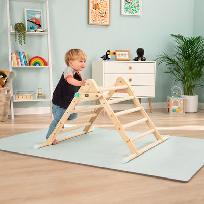 A toddler stands and plays on the TP Toys Energetic Play Pack: TP Active-Tots Pikler Style Wooden Climbing Triangle in a bright, well-decorated children’s room filled with toys and plants.