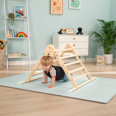A young child crawls under the TP Toys Energetic Play Pack: TP Active-Tots Pikler Style Wooden Climbing Triangle with Owl & Fox Hammer Bench and Xylophone on a play mat in a light blue, neatly decorated playroom.