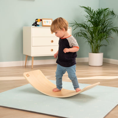 A young child stands barefoot on the TP Toys Active-Tots Pikler Style Climbing Triangle, Bridge, Climbing Cube & Balance Board—FSC® certified—in a playroom with light blue walls, a potted plant, and a white dresser.